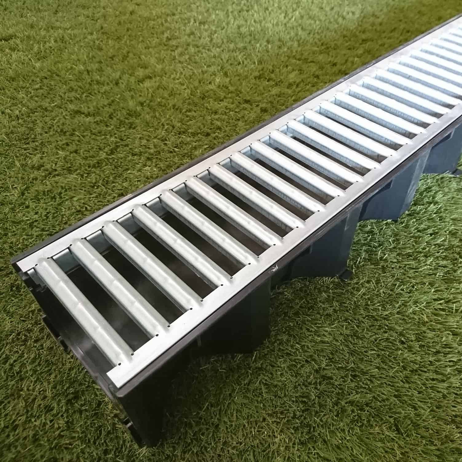 The ACO Galvanised Drain Unit, featuring a slotted grate with parallel bars for efficient water drainage, is placed on green artificial grass.