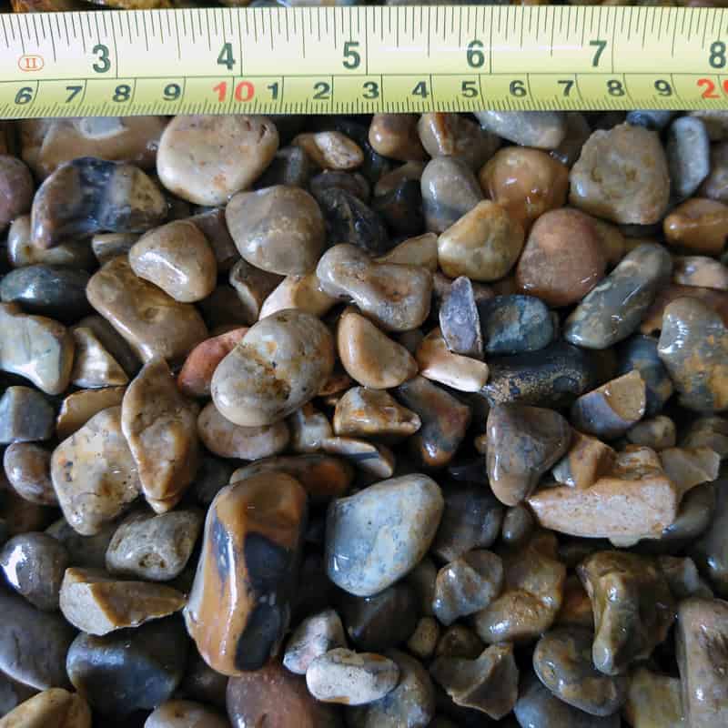 Displayed next to a measuring tape, the Natural Rounded 20mm Golden Pebbles show their smooth, multicolored variety in hues of brown, gray, and beige. The measuring tape highlights that some pebbles measure precisely 20mm for size comparison.