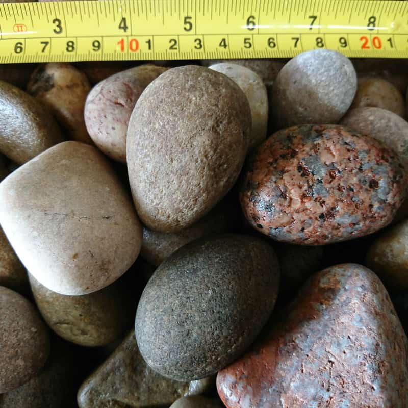 A measuring tape in centimeters above smooth, oval-shaped Scottish Highland Pebbles shows their size range of 30-50mm; these bagged stones vary in multicolored hues of brown, gray, and red.