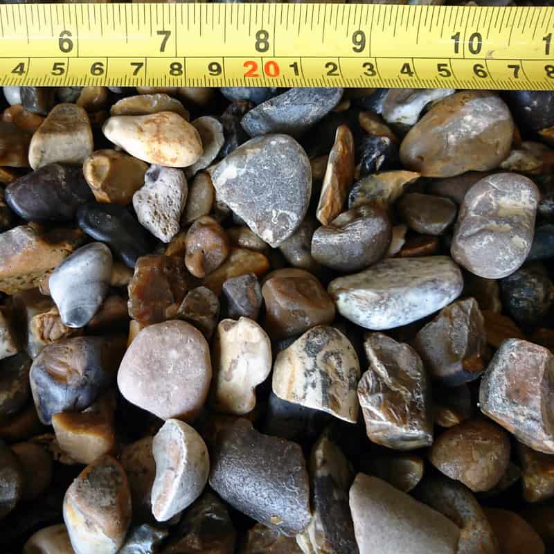 A close-up of natural mixed 10-20mm Blue/Grey Gold Pebbles shows brown, gray, and white tones with a yellow measuring tape indicating sizes from 5 to 20 cm.