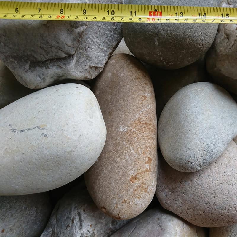 A close-up of 100-200mm Highland Glacial Cobbles in gray and brown, with a yellow measuring tape above showing measurements in centimeters. The longest stone is approximately 18 cm.