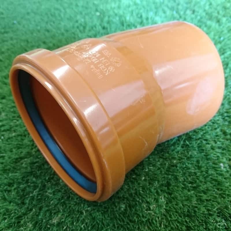 The 110mm Single Socket Bend, a brown PVC pipe connector with a rubber seal, is on the green grass. It's cylindrical with one end slightly larger for smoothly joining pipes at a gentle 15° angle.