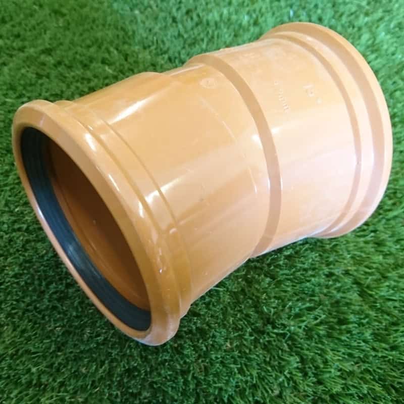 A 110mm 15° Double Socket Bend, featuring a brown ridged design and black rubber rings inside, rests on grass. The cylindrical connector is angled at 15° and made of a plastic-like material.