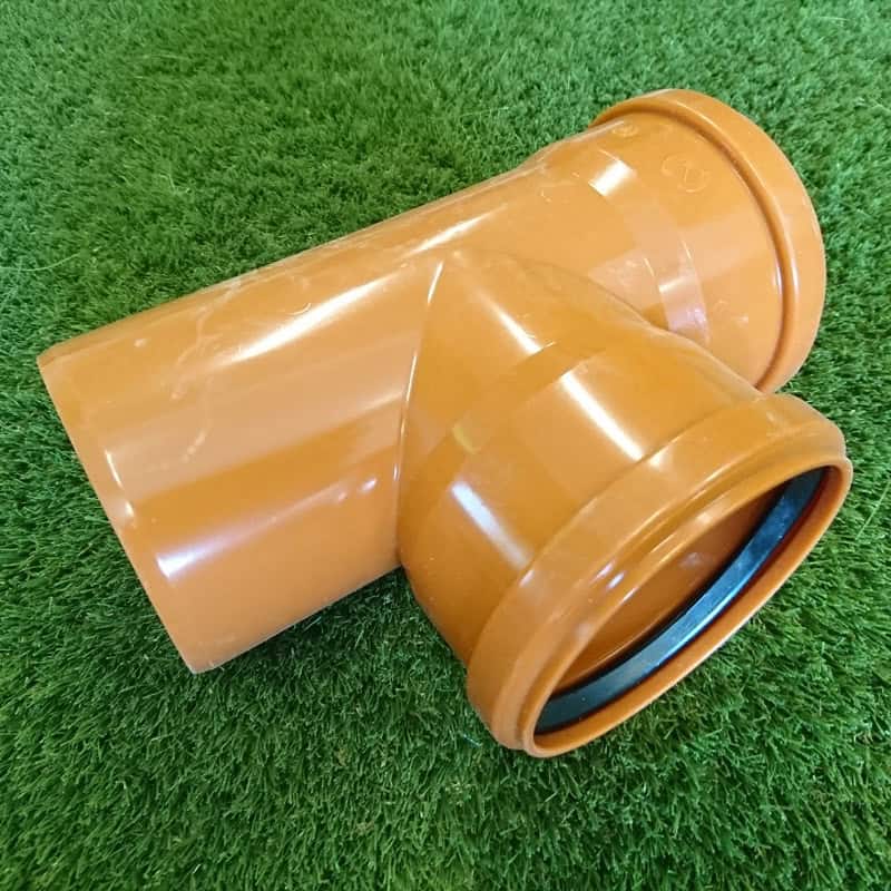 A 110mm 90° Double Socket Junction in orange PVC, designed for plumbing or drainage connections, rests on green artificial grass. This T-shaped fitting features smooth surfaces ideal for connecting pipes.