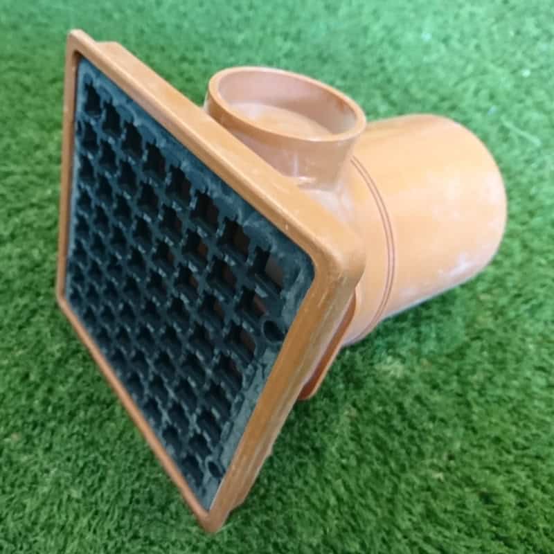 A 110mm Square Hopper Spigot End, featuring a brown drainage pipe and a black, gridded cover of small squares, rests seamlessly on green grass.