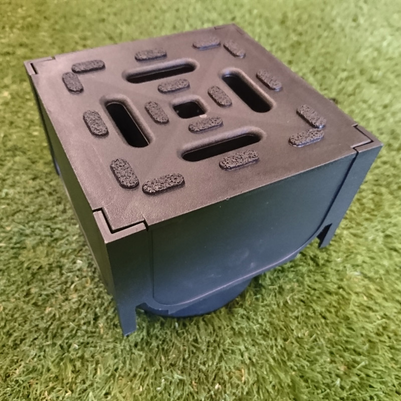 A black ACO Drain Corner Unit, featuring a square design with top vent slots, sturdy corner supports, and a central circular opening at the bottom, is placed on artificial grass.