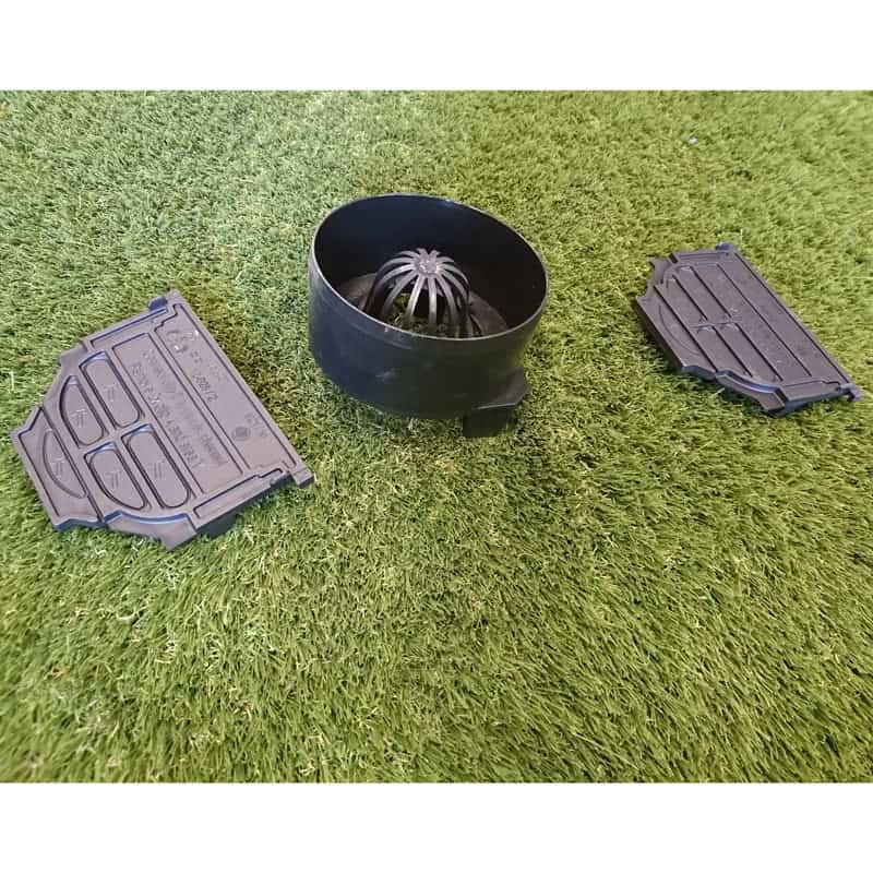 The ACO Drain Accessory Kit is displayed on artificial grass, showcasing a central black cylindrical piece with a domed grate and two rectangular side pieces with slotted designs for improved functionality and aesthetic appeal.