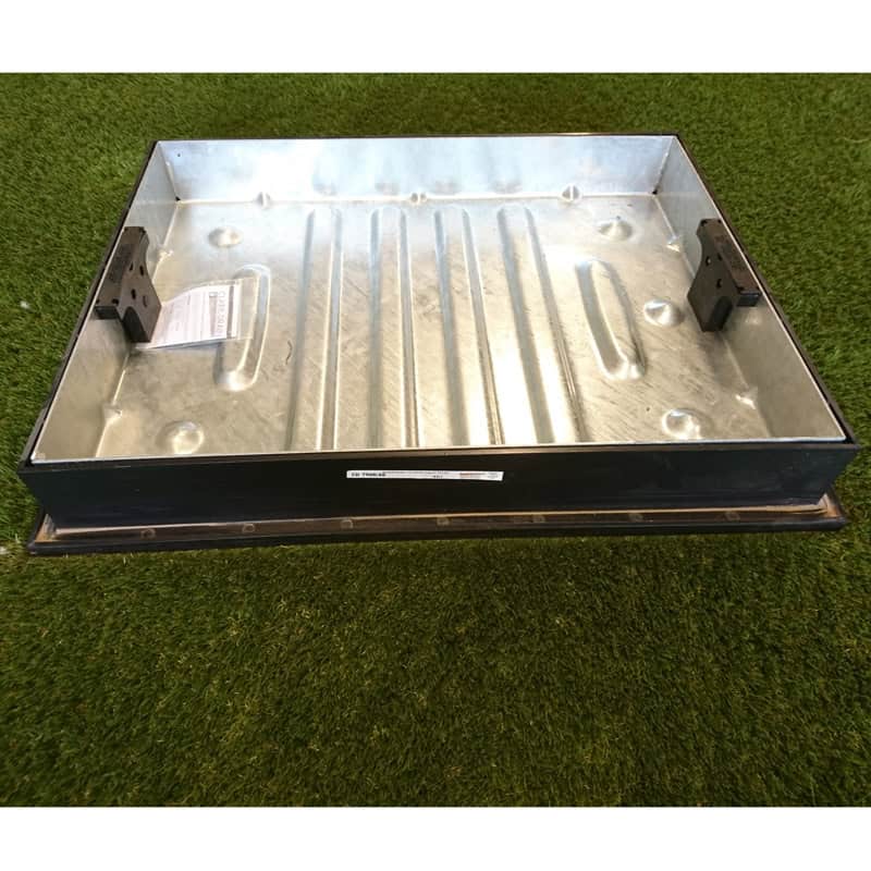 A 600x450x80mm Recess Cover & Frame with raised edges and brackets on opposite sides rests on green turf, holding a piece of paper. The surface has a ribbed texture.