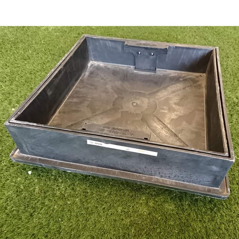 A 320mm Round to Square Recess Frame & Cover rests on artificial grass, showcasing modern design with functionality. The black square manhole cover and open frame feature a small top handle and a light-colored side label.