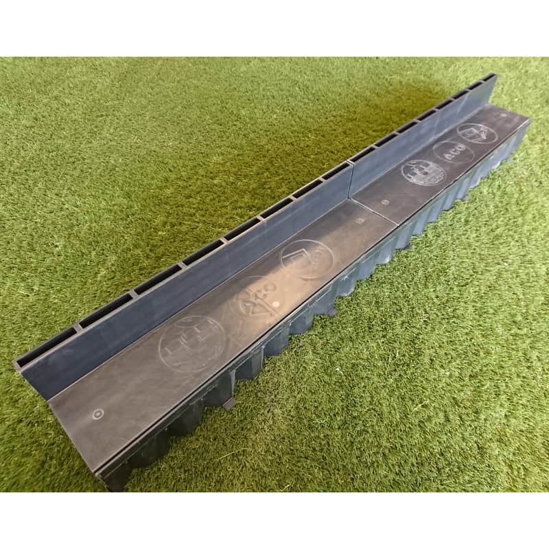 The ACO Concealed Drain Unit, a black plastic drainage channel, is installed on artificial grass and features vertical grooves and embossed symbols on its surface.