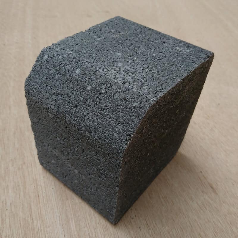 The Barleystone Paving 2 Way Small Kerb, a textured dark gray concrete brick with a curved side, is placed on a light wooden surface. Its rough surface and unique shape are clearly visible.