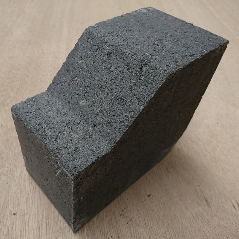 A large, gray Cemex 2 Way Large Kerb High block with an irregular shape and wavy top surface sits on a wooden table, resembling a rough piece of kerb stone.