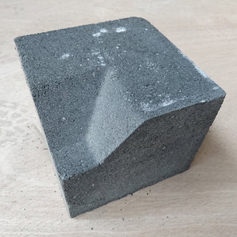 A close-up of a gray concrete block with a smooth, angled cut on one side resembles the Barleystone Paving 3 Way Large Internal Corner Low. It rests on a light wooden surface, showcasing its texture and minor surface imperfections.