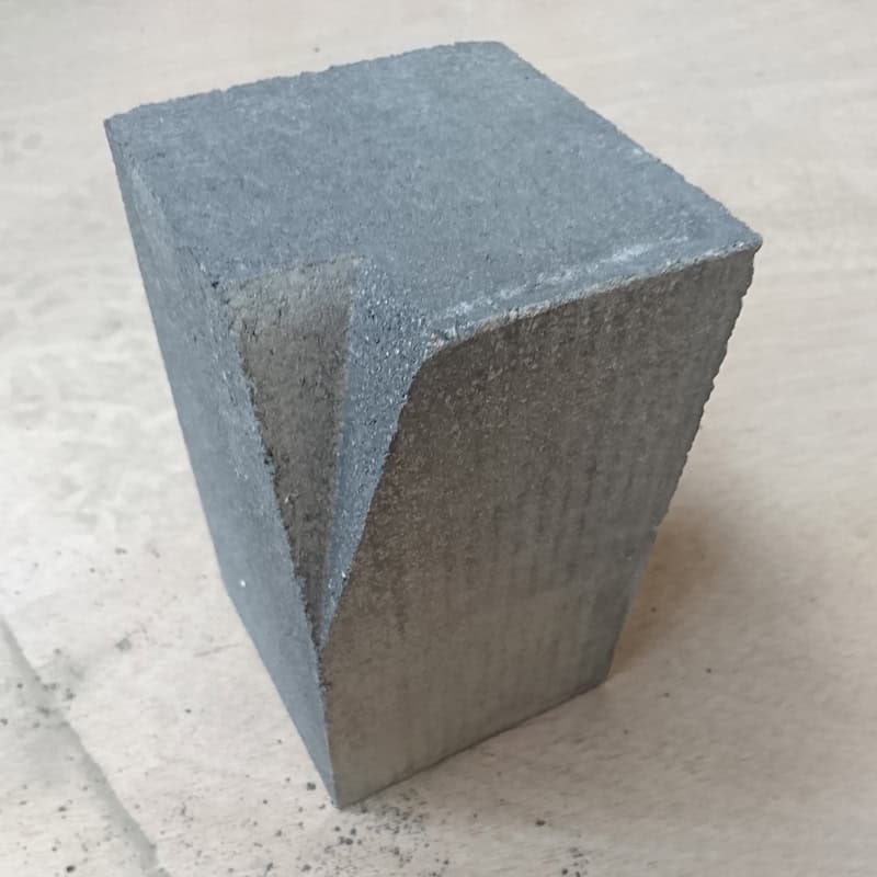 A rectangular concrete Barleystone Paving 3 Way Large Internal Corner High block stands on a light surface, featuring a triangular cutout with a rough texture. Dust surrounds its base, adding mystery and intrigue.