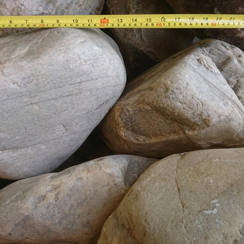 A close-up image displays 400-600mm Highland Glacial Boulders with a yellow tape measure, marked in centimeters, stretched across them to highlight their impressive size. The boulders feature a blend of light brown and gray hues.