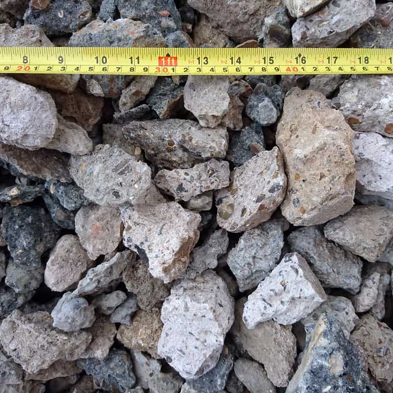 The pile of Crushed Concrete - Loose consists of grey and beige construction rubble with rocks of various sizes. A yellow measuring tape, marked in inches, stretches across the heap and shows measurements up to 50 inches.