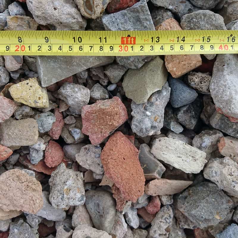 A close-up image of Crushed Brick Rubble - Loose in red, gray, and beige hues is mixed with loose debris. A yellow tape measure runs across the top, showing measurements in centimeters and inches.