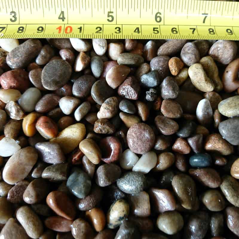 A scattering of small, smooth multicolored Scottish Highland Pebbles (08-14mm) is shown beneath a yellow measuring tape with black and red markings up to 9 cm, perfectly illustrating the size range of the bagged pebbles.