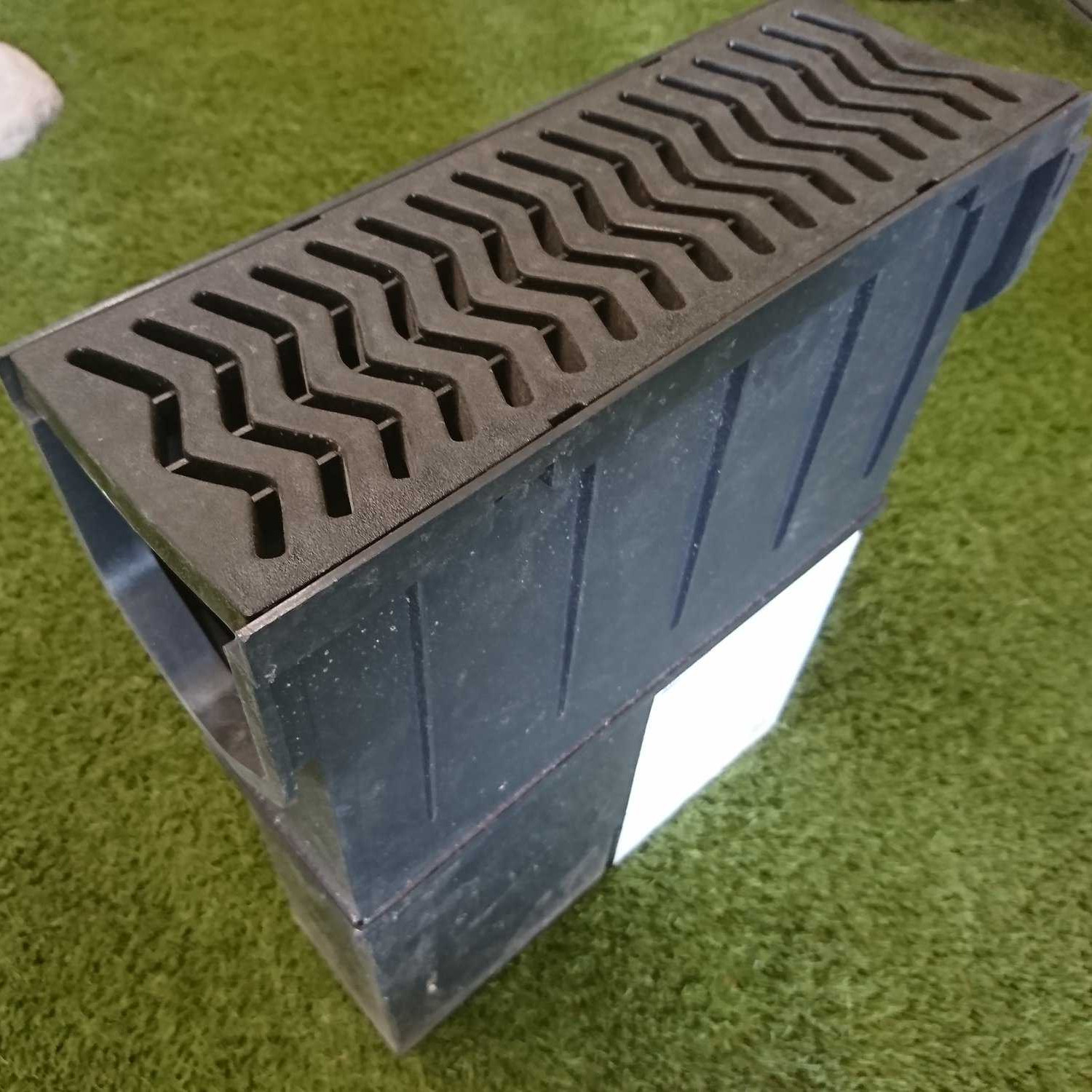 On lush green artificial grass, the FERNCO black rectangular drainage channel, featuring a zigzag-patterned grate cover and the optional FERNCO Sump Unit, offers efficient water management and integrates seamlessly into any landscape.