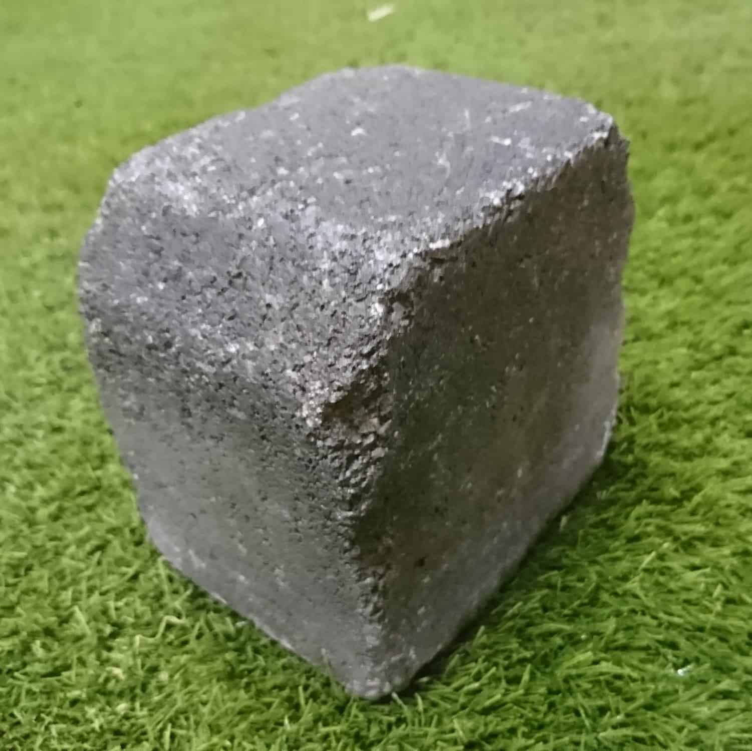 A rough, gray concrete cube resembling the Barleystone Paving 2 Way Cobble Small Kerb rests on a patch of green grass, showcasing a solid and textured appearance that contrasts with the vibrant grass.