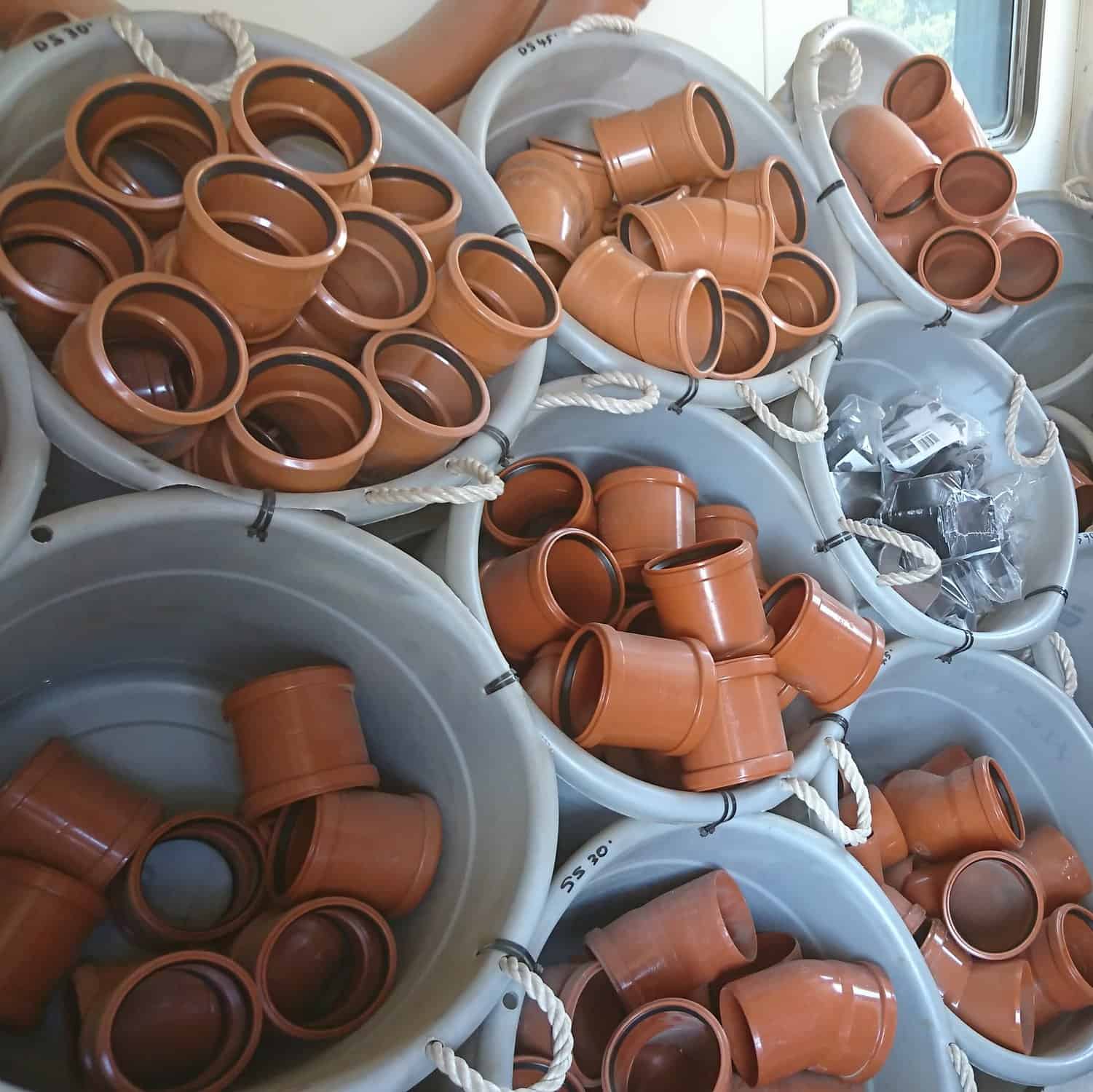 Several large white tubs, each with rope handles, are filled with 110mm Double Socket Slip Couplers and some metal parts. The tubs are stacked tightly together.