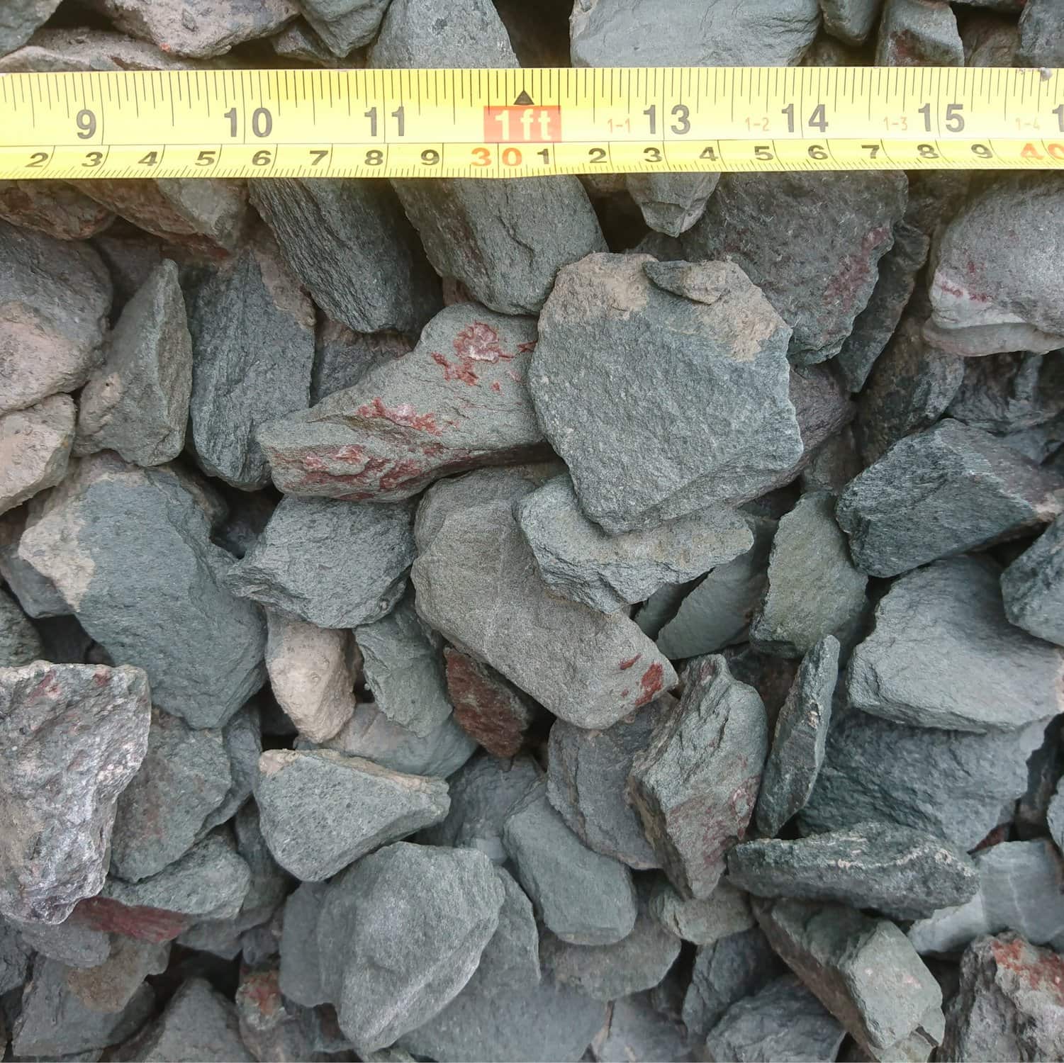 Close-up of several Slate Green Grey 40mm rocks with a yellow measuring tape above them showing just over 1 foot in feet and inches. Some rocks have reddish markings that contrast with their green-grey hues.