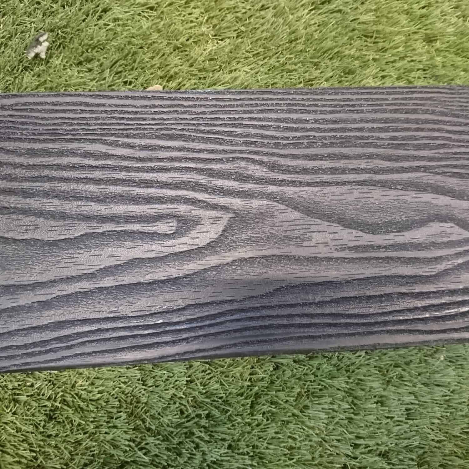 A close-up of the Composite Decking Anthracite Black showcases its textured, wood-like surface with visible grain patterns, set against a vibrant green grass backdrop.