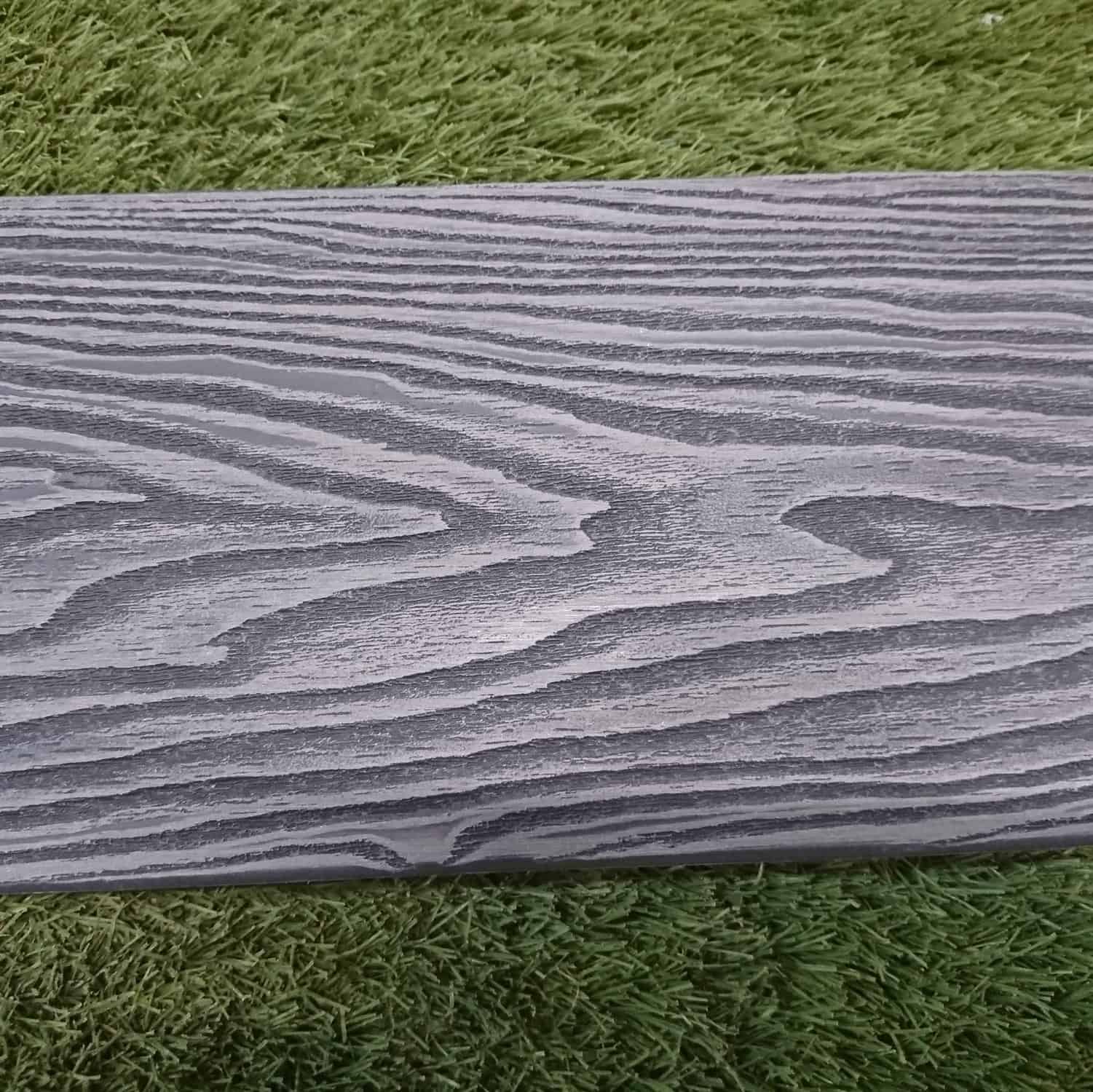 A close-up of Composite Decking in Mid Grey, featuring a textured pattern similar to a rectangular wooden plank, rests on bright green grass.