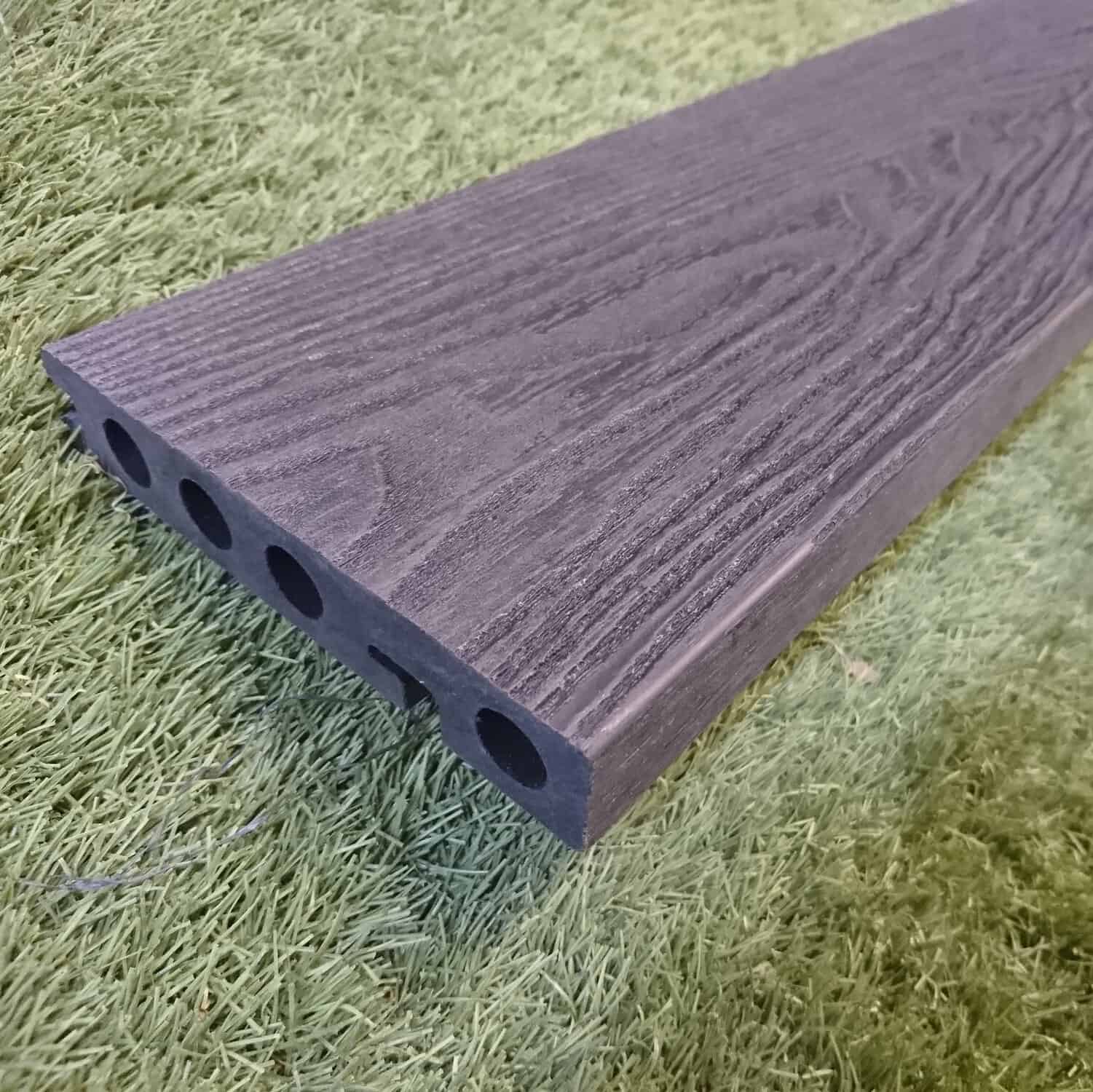 Close-up of a Composite Decking Edging Board in anthracite black with wood grain texture and four hollow channels, resting on green artificial grass.