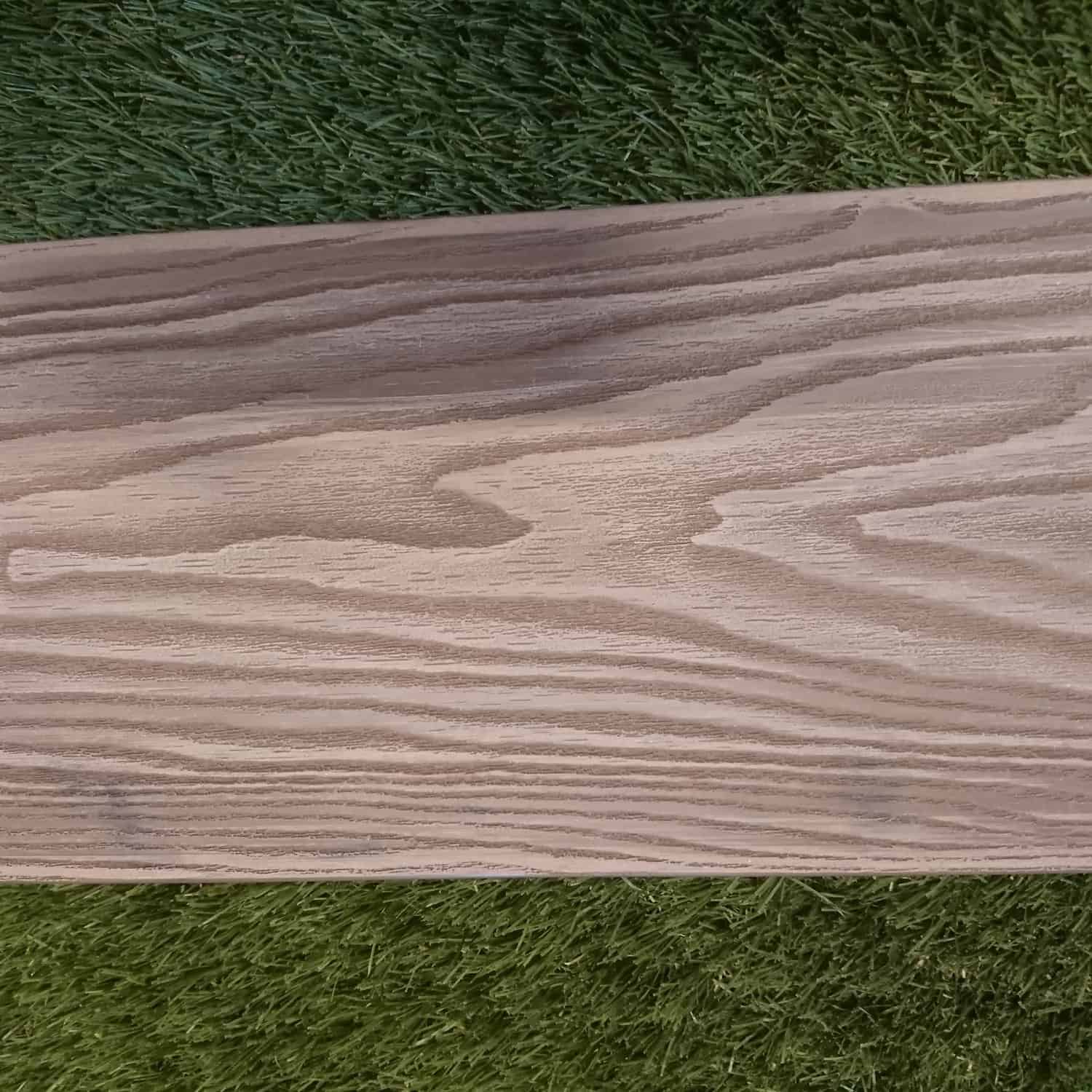 The Composite Decking Brown plank, featuring visible grain patterns, lies on green artificial grass. This pairing juxtaposes the natural wood texture with synthetic grass for a striking contrast.