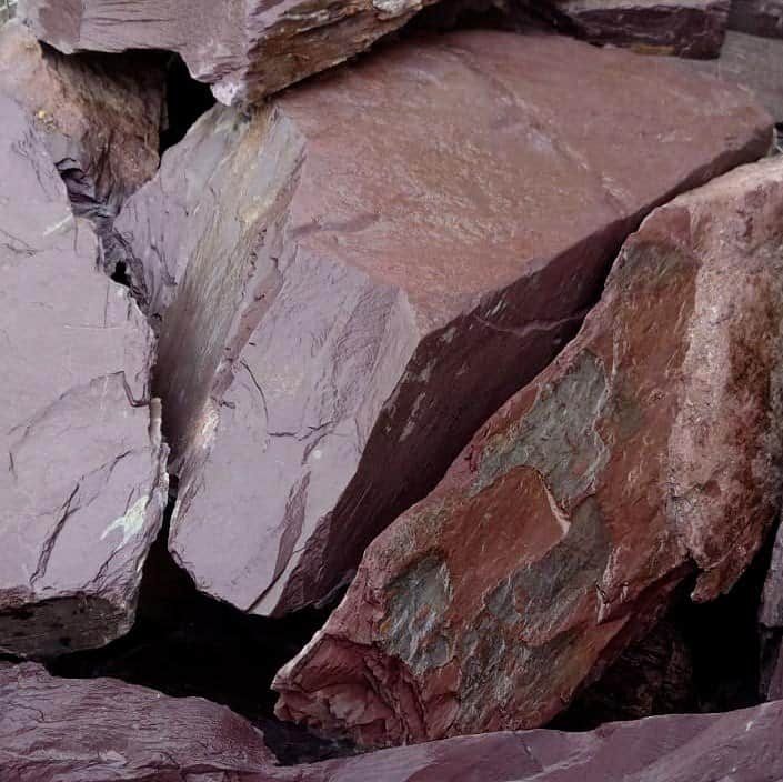 A close-up of several large, jagged rocks from the 300-400mm Plum Slate Rockery displays a blend of earthy colors including purple, gray, and brown. The rocks have rough, uneven surfaces and are arranged in a natural rockery design.