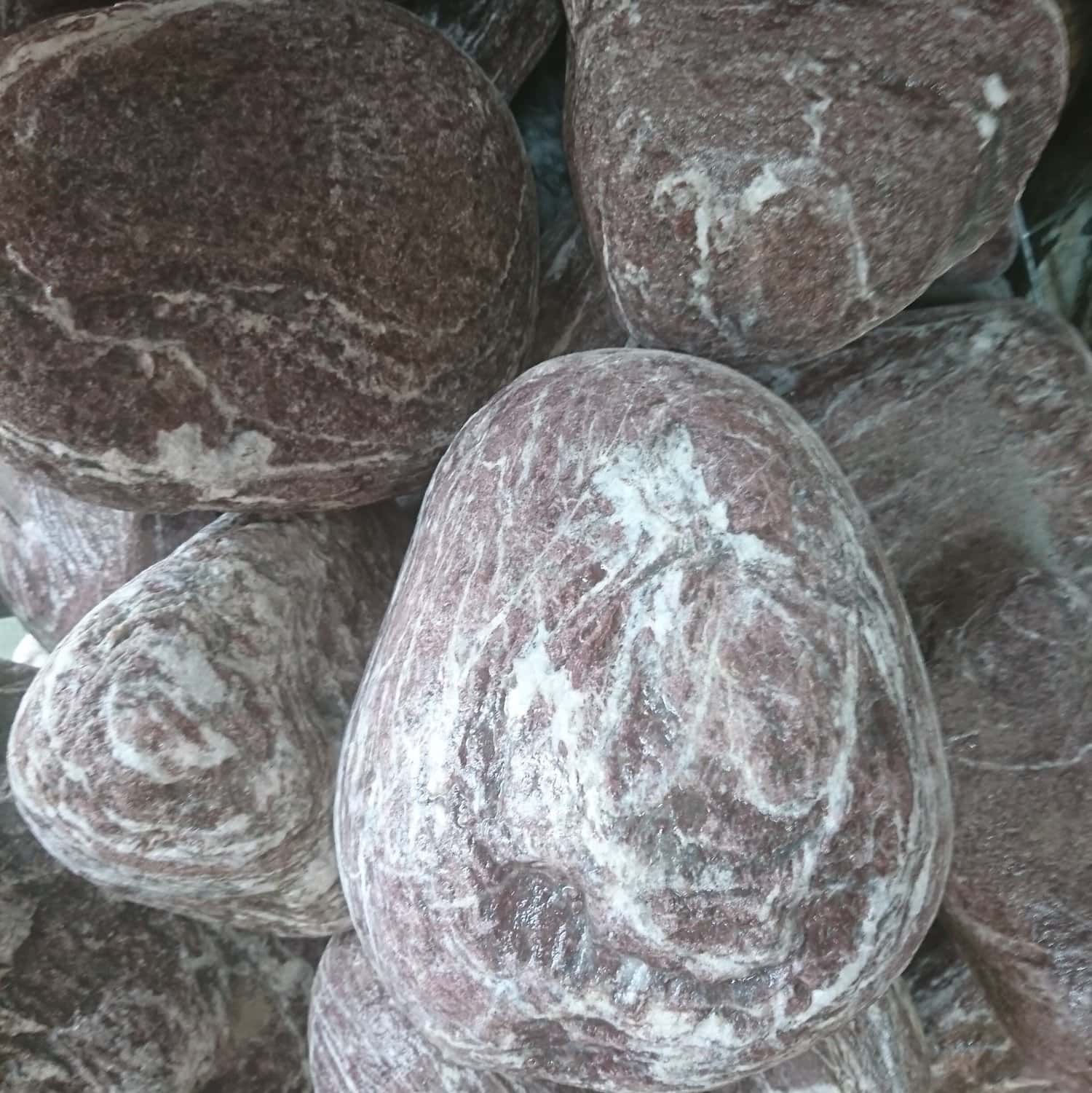 The 200-300 Leylac Marbled Boulders are smooth, oval-shaped rocks with dark reddish-brown hues and white marbled patterns. They are closely arranged, highlighting their glossy surfaces and unique textures.