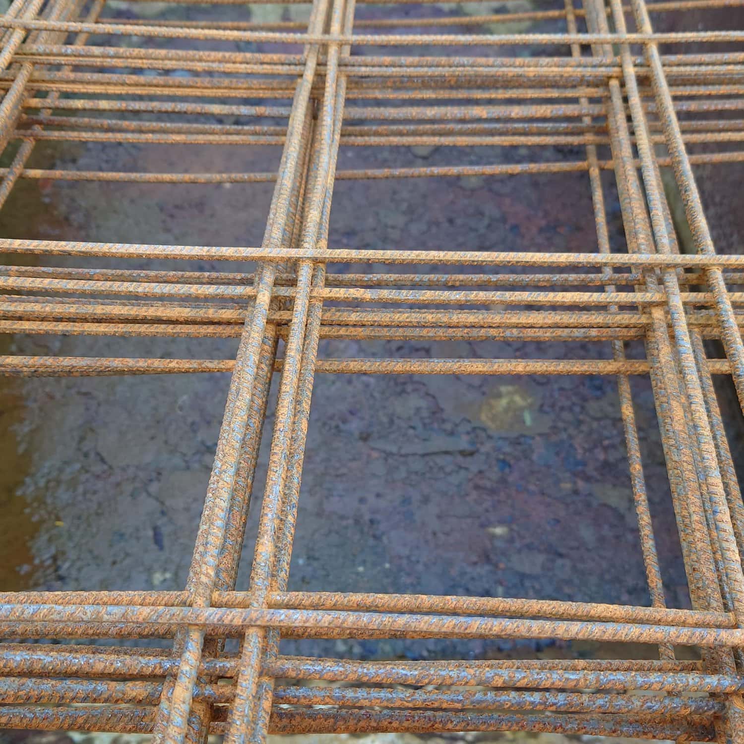 Close-up of a Steel Mesh A142 (2.4m x 2.4m) grid used in construction, featuring a crisscross pattern over rough concrete; the steel bars exhibit a weathered texture with brownish-orange rust.