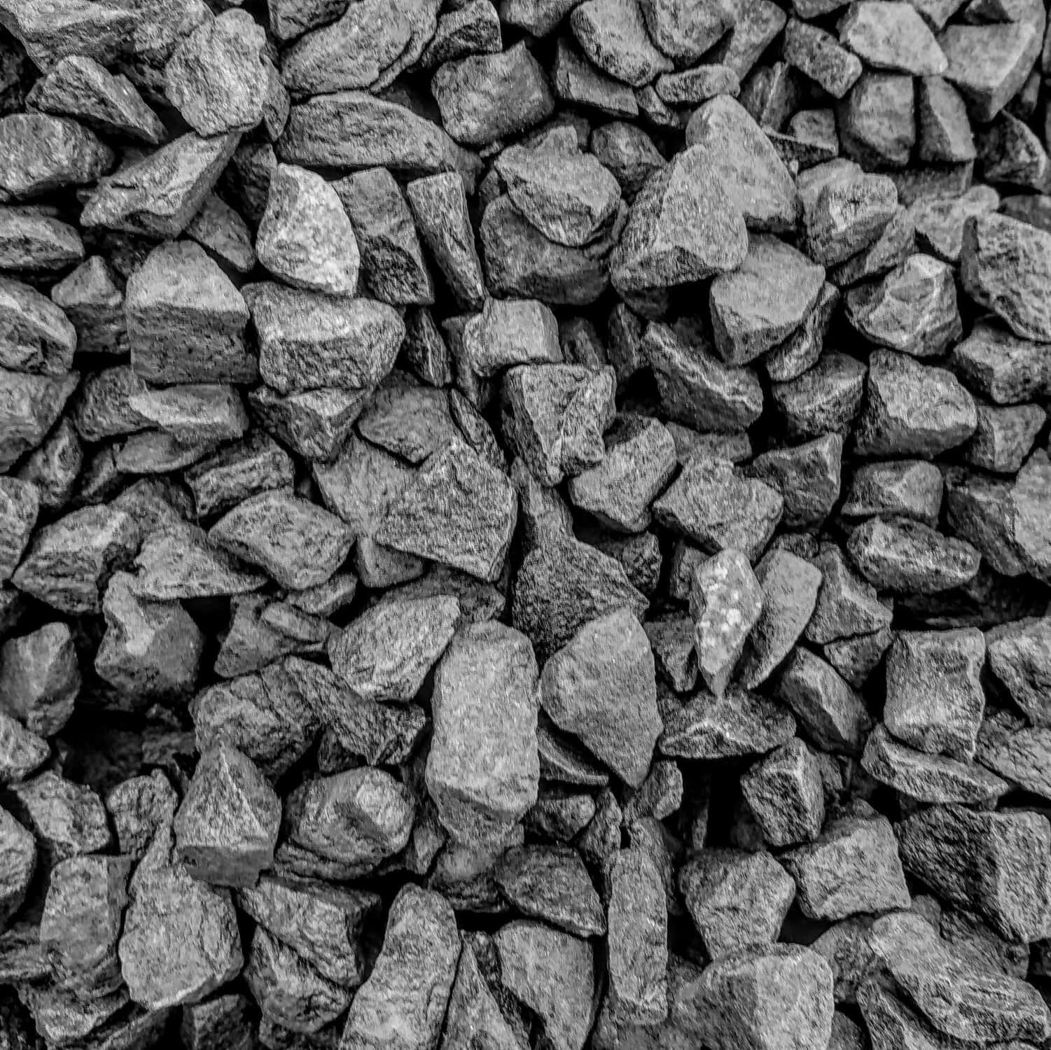 A close-up showcases a dense pile of Black Basalt 14-20mm Chippings - Loose, featuring irregular, rough stones in shades of gray. The varied-sized chippings fill the entire frame, creating a rugged appearance.