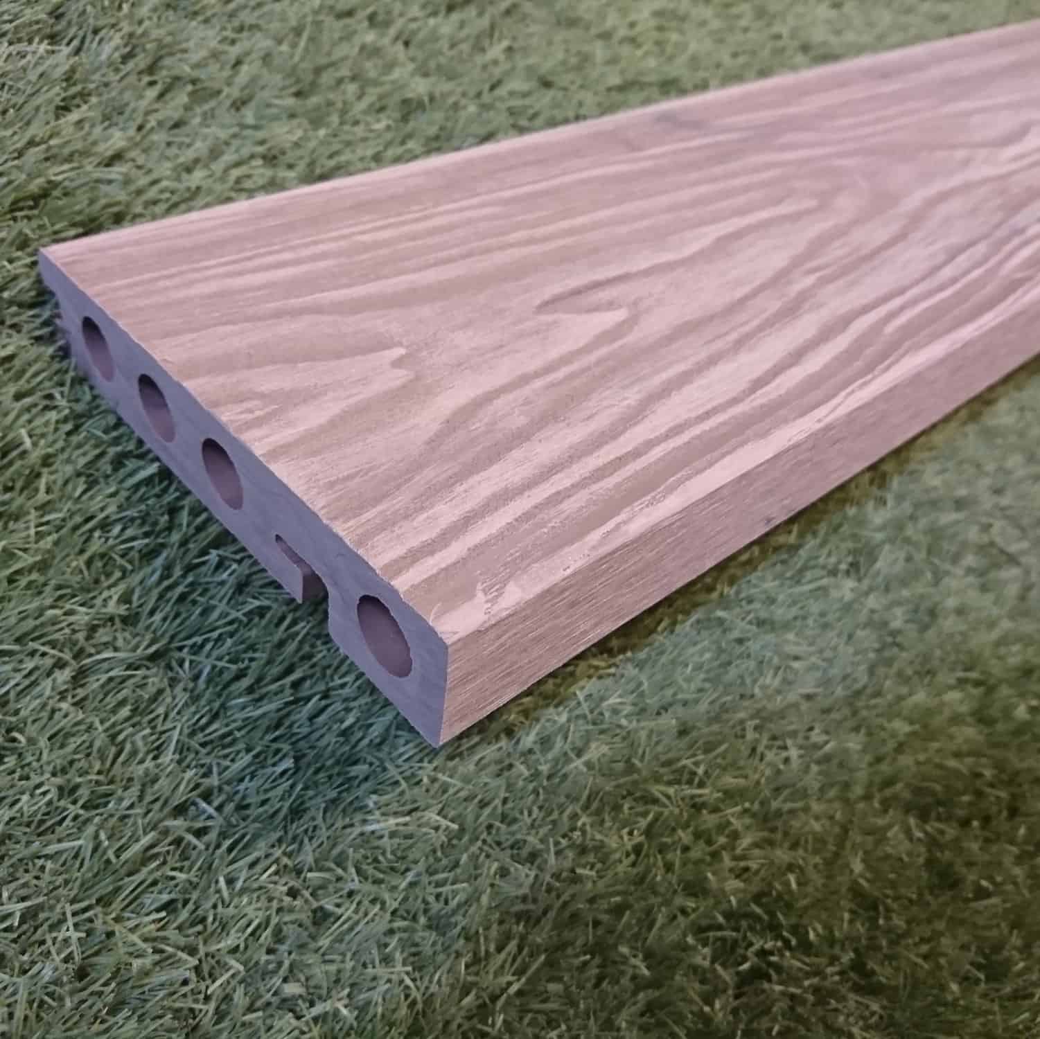 The Composite Decking Edging Board Brown, with woodgrain texture and circular hollow sections for a lightweight design, elegantly rests on a green grass surface.