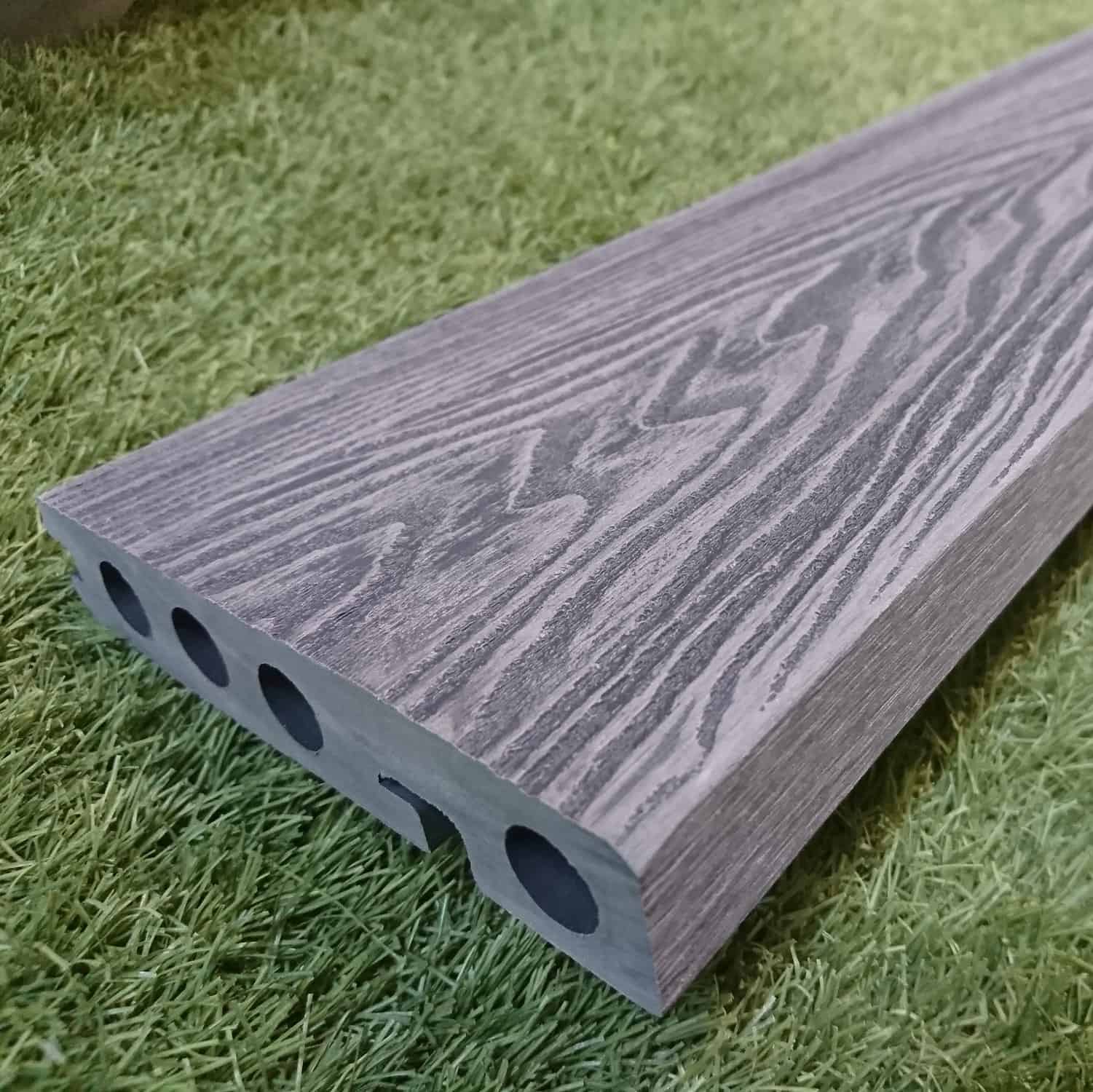 A close-up of the Composite Decking Edging Board in mid grey showcases its wood grain texture and side holes, resting on lush green artificial grass.