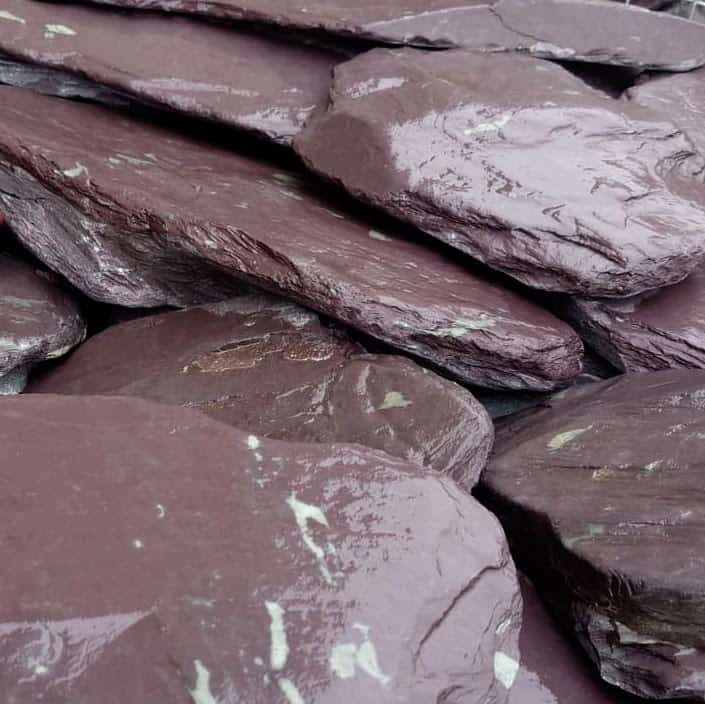 A close-up of stacked 200-300mm Celtic Slate Paddle/Stepping Stones showcases dark maroon hues, smooth texture, reflective spots, and subtle white markings.