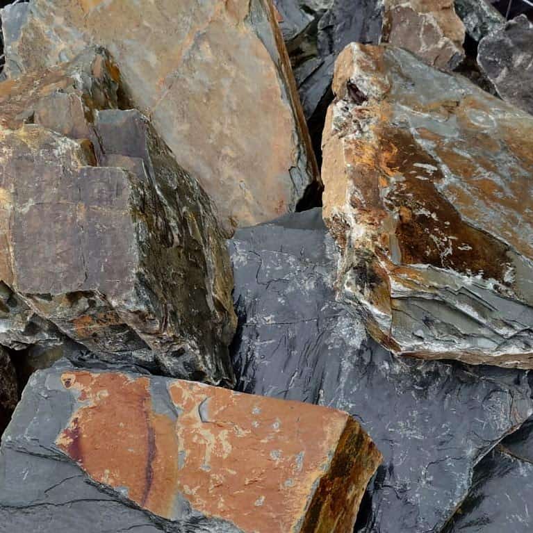 A close-up view of the 300-400mm Rustic Slate Rockery reveals a collection of slate rocks in earth tones like grays, browns, and rusty orange, featuring rough, jagged surfaces and layered textures.