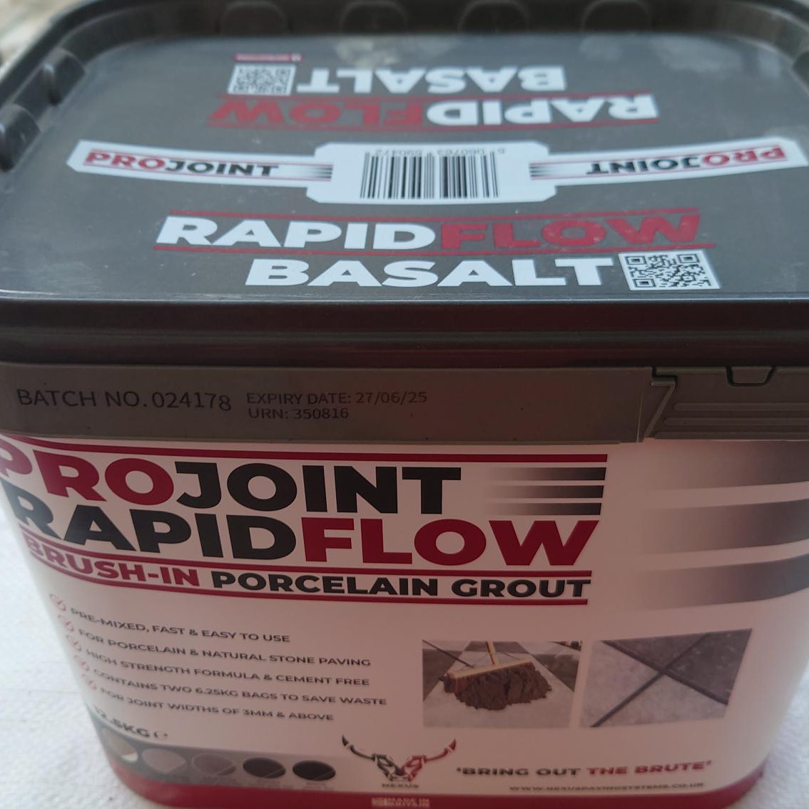 A rectangular container of ProJoint Rapidflow PORCELAIN Grout (Basalt) is on a white surface. The lid showcases product branding and a QR code, with part of the bag visible in the background.