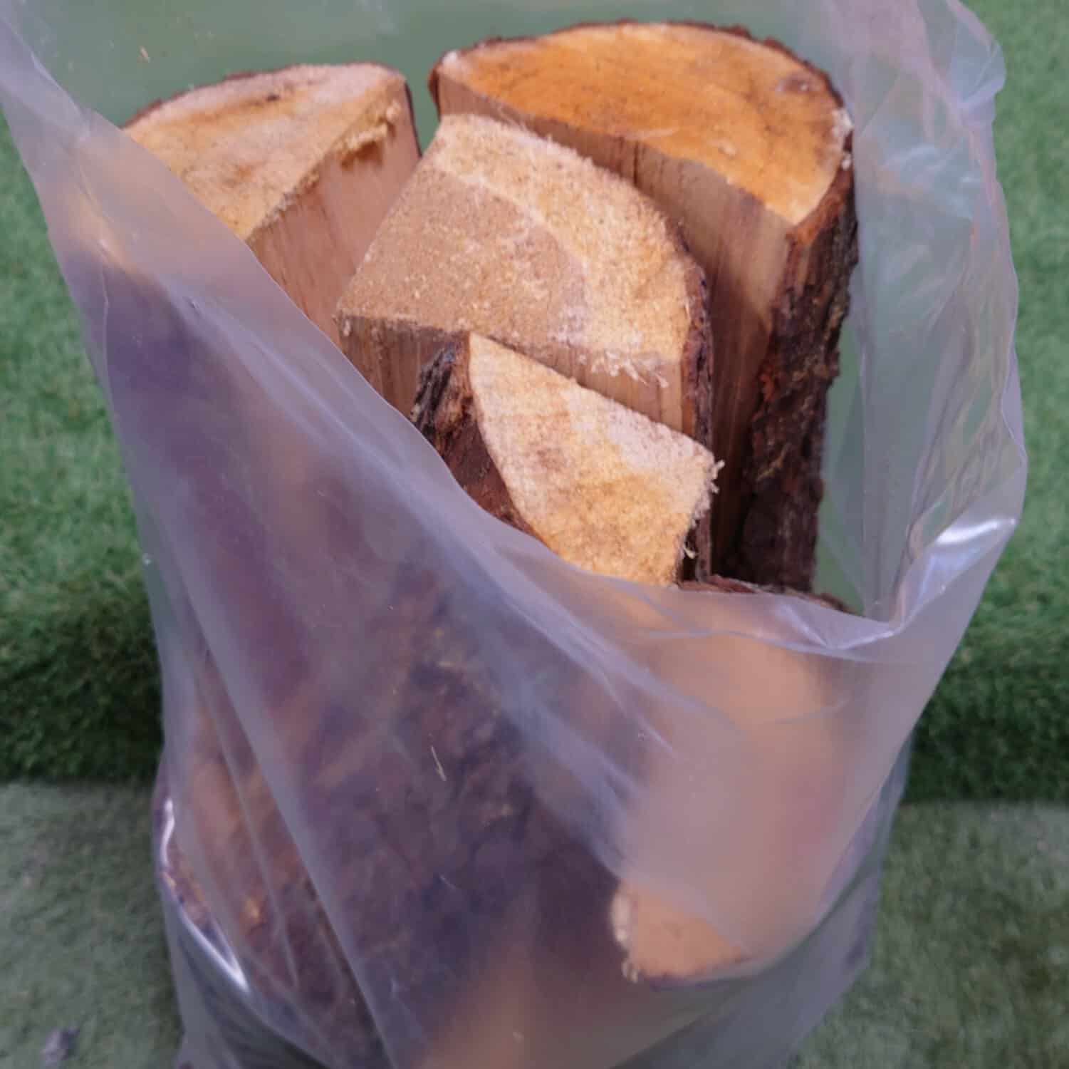 The Kiln Dry Hard Wood Logs Handy Bag contains 10 logs with smooth surfaces and intact bark, all encased in a transparent plastic bag on a grass-like surface.