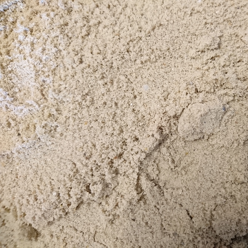 A close-up shows a pile of Building Sand - Loose, featuring a beige color and slightly coarse texture akin to fine flour or sand. The uneven surface displays small clumps and shade variations.