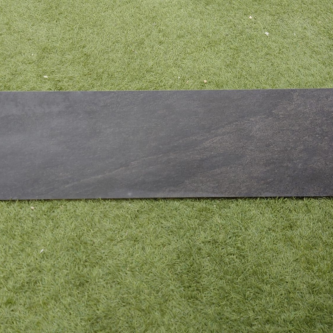 A Premier Cathedral Anthracite Porcelain Edge Boarder, sized 900x200x20mm, lies horizontally at the center of a lush green grass surface.