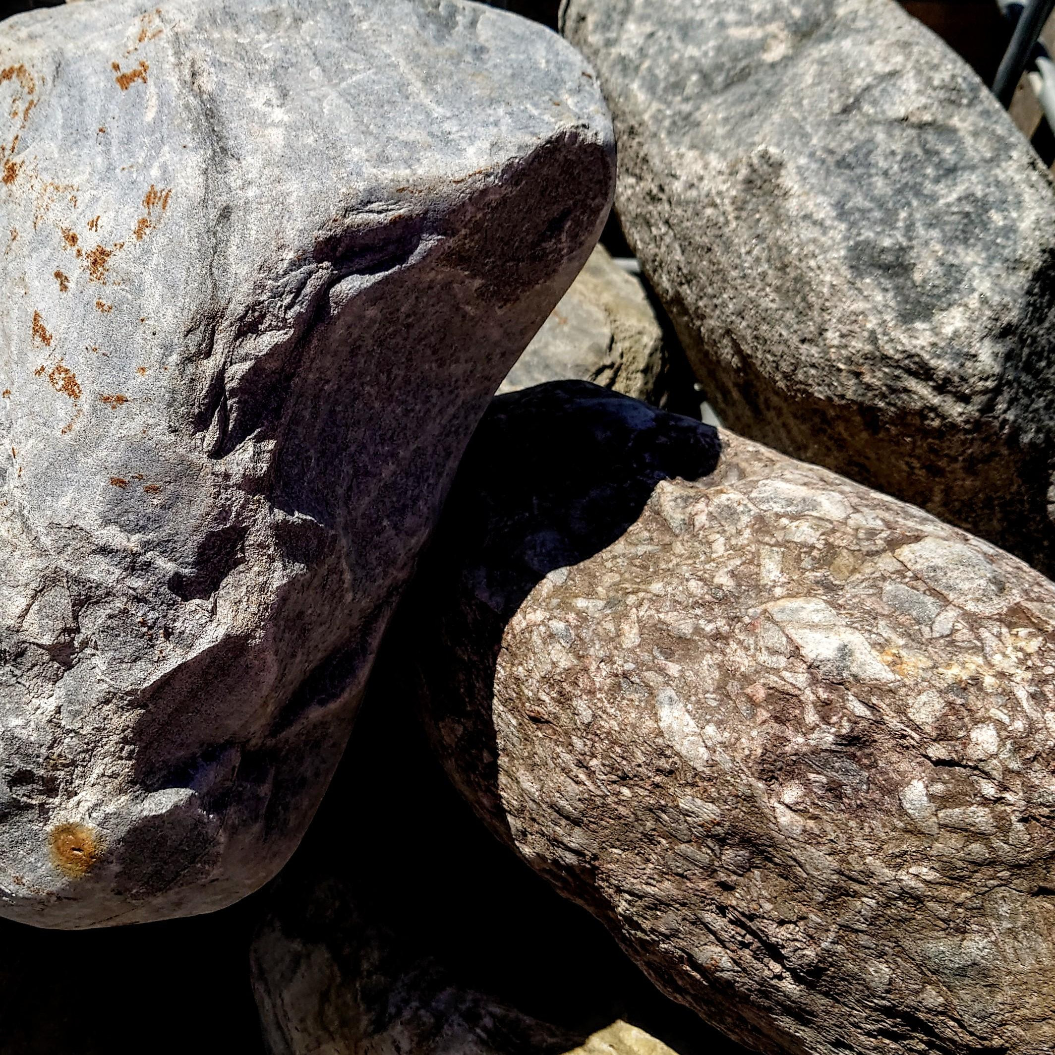 A pile of 500-800mm Highland Glacial Boulders, showcasing large, textured surfaces in gray and brown shades with irregular patterns and hints of orange and white, catch the sunlight, casting intricate shadows.