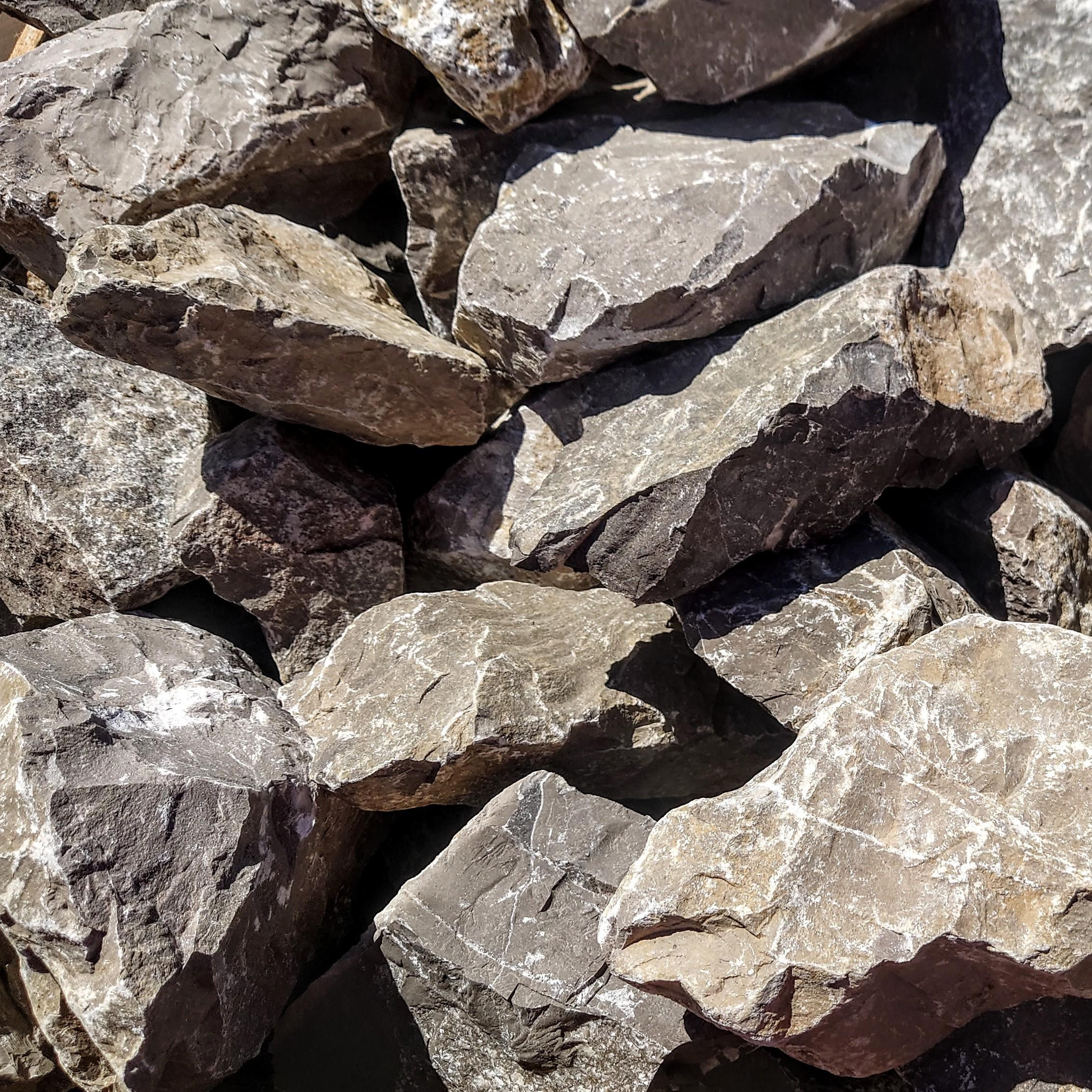 A close-up of a pile of irregularly shaped 300-400mm Longstone Rockery pieces, showcasing their gray and brown rough, textured surfaces. Sunlight casts shadows, emphasizing the jagged edges and crevices.