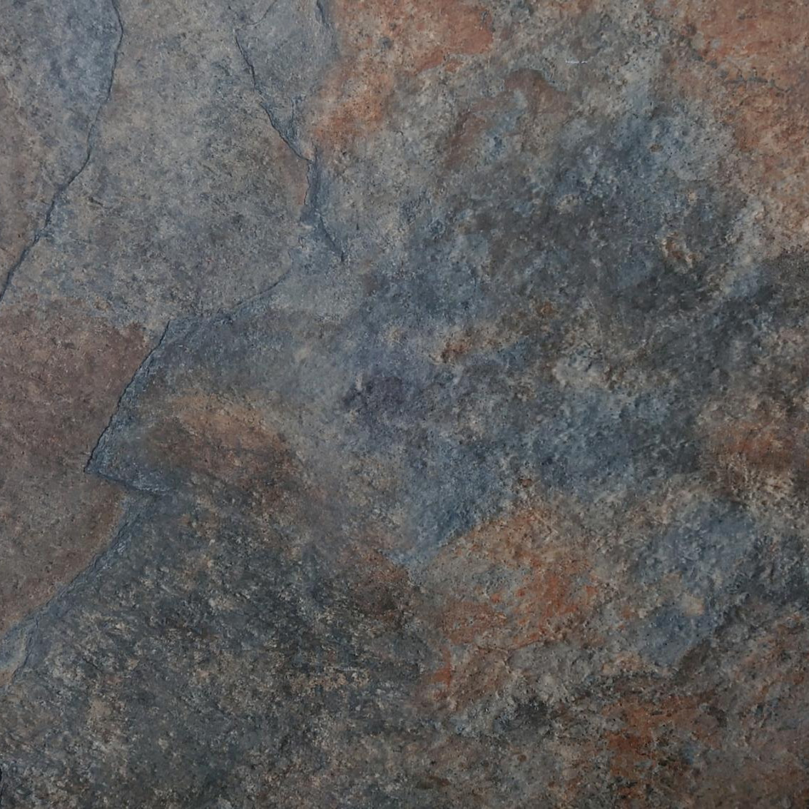 A close-up of Premium Rustic Slate Porcelain 900x600x20mm showcases a textured stone surface with blue, brown, and gray hues, visible cracks, and natural patterns. Its rugged look and premium porcelain finish evoke timeless elegance.