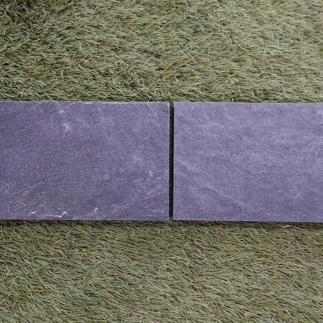 Two Nuova Anthracite Porcelain Edge strips, 100x200x20mm, lie side by side on a textured green surface resembling artificial grass, revealing subtle color variations against the vibrant backdrop.