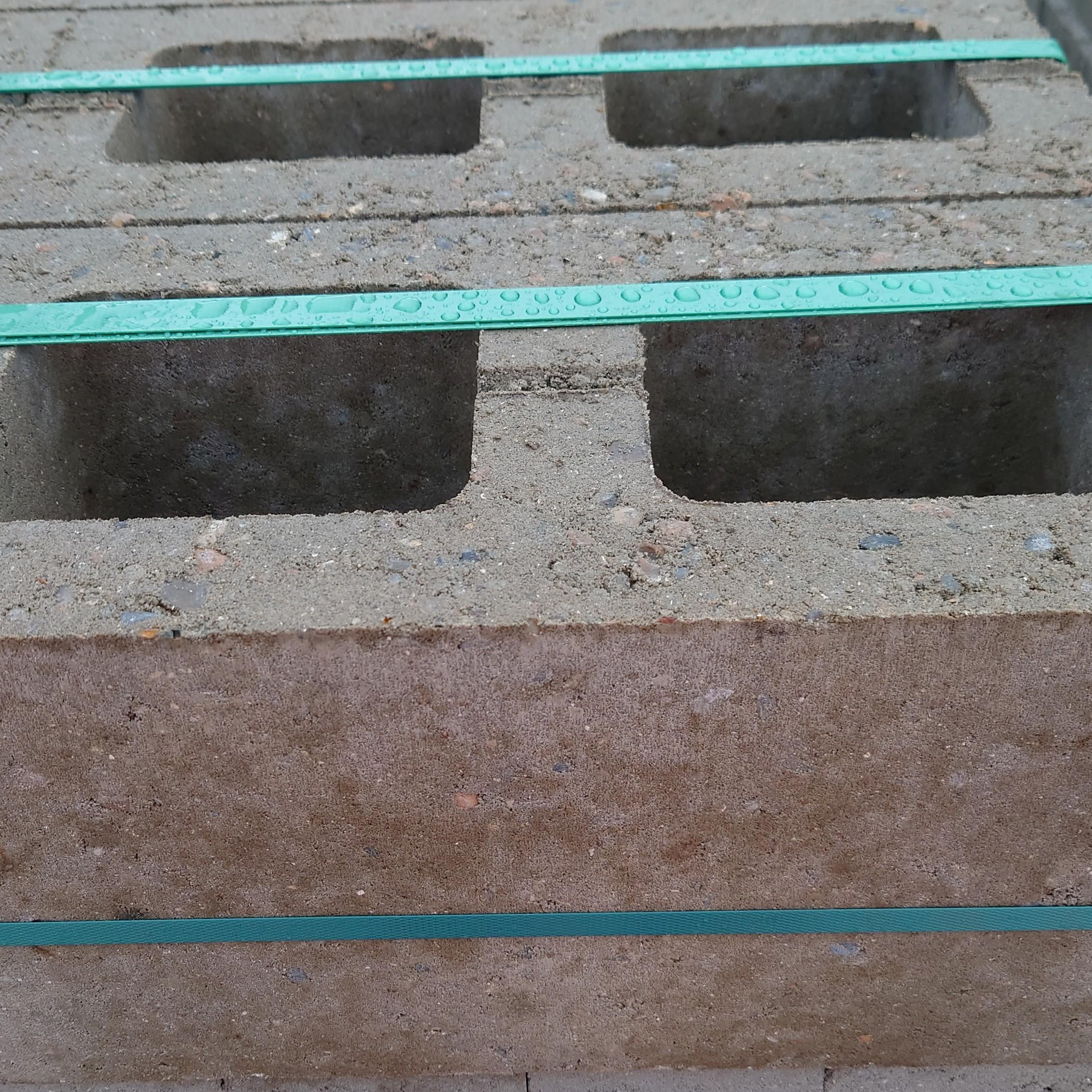 Close-up of CEMEX (Large) Hollow Concrete Blocks 7.3 n featuring rectangular holes, stacked neatly. Two green measuring tapes lie across the top, highlighting scale and alignment with the rough texture and grainy surface clearly visible.