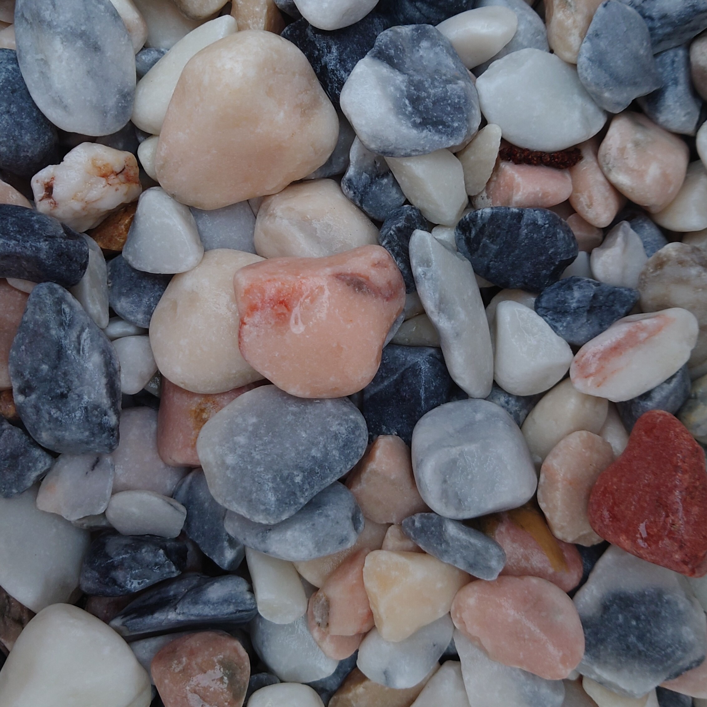 A close-up of smooth, rounded stones reveals a blend of blue, gray, pink, and white hues. Some are glossy while others are matte, resembling the diverse Flamingo 20-50mm Pebbles - Bagged collection.